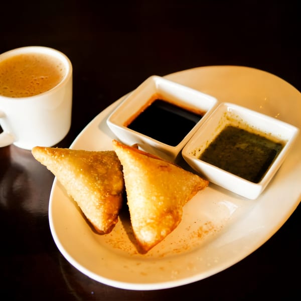 Two golden-brown samosas with two dipping sauces and a cup of chai on a white plate.